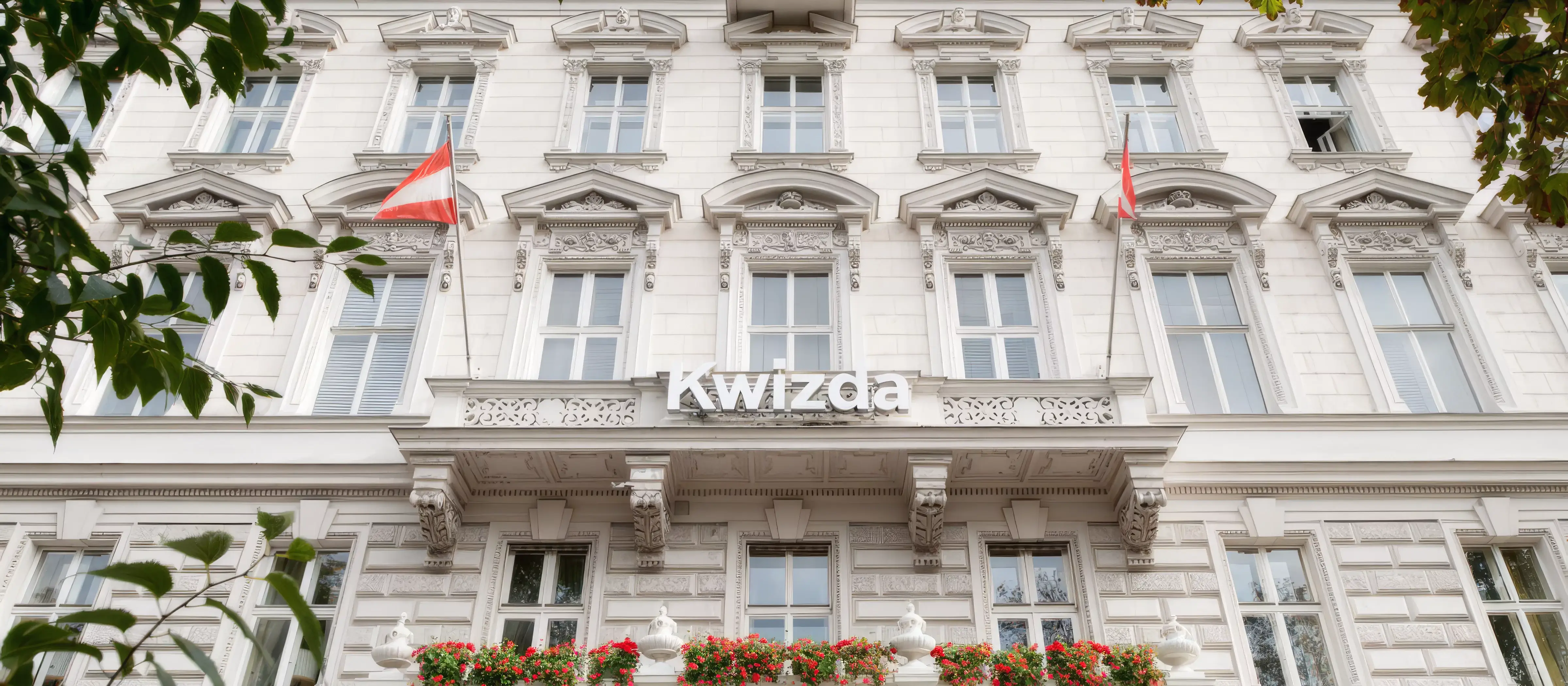 Fassade eines historischen Gebäudes mit dem Schriftzug "Kwizda", verziert mit Balkonen, roten Blumen und zwei Flaggen an der Front.